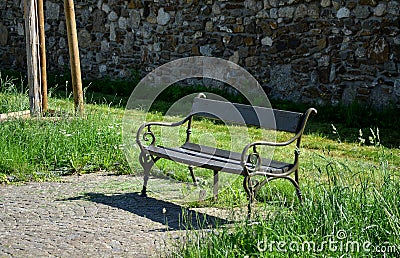 Stone wall in front of it stands a bench of metal twisted pipes and wood on a paved stone path meadow with flowers in a small town Stock Photo