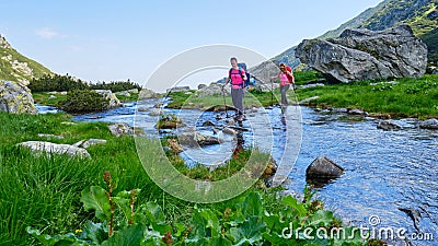 Two active, fit women hikers crossing a mountain river by stepping on rocks, with heavy camping backpacks and trekking poles. Stock Photo