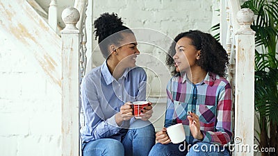 Two african american curly girls sistres sitting on stairs have fun laughing and chatting together at home Stock Photo