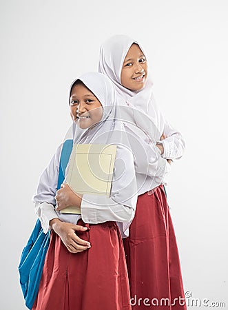 two Asian veiled girls wearing elementary school uniforms stand back to each other while carrying a backpack and a book Stock Photo