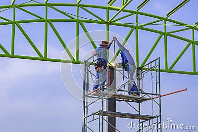 Two Asian welders on scaffolding are welding metal roof structure of industrial building against blue sky Editorial Stock Photo