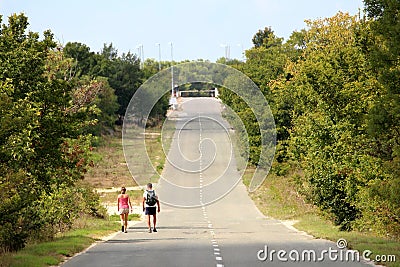 Two backpacker tourists walking on long straight paved road going uphill towards closed site with ramp surrounded with dense Editorial Stock Photo