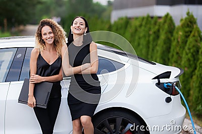 Two beautiful girls in black outfut smiling and posing. Businesswomen with laptop and electric car with charging socket plugged in Stock Photo