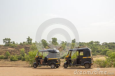 A two black and yellow Indian motor rickshaws stand on an asphalt road against the background of a hill and green forest Stock Photo