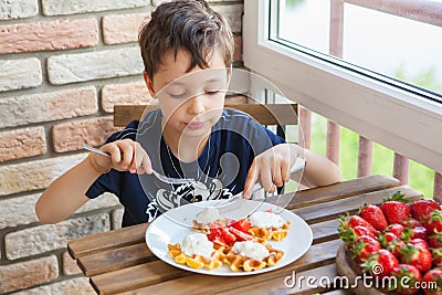 Two boys are eating waffles with strawberries and ice-cream over a wooden table on the balcony Stock Photo