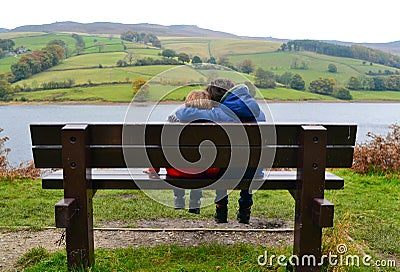 Two brothers sitting on bench with older brother cuddling younger sibling. Overlooking river and hills. Stock Photo