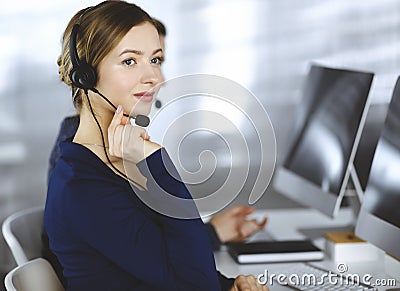 Two busineswomen have conversations with the clients by headsets, while sitting at the desk in a modern office. Diverse Stock Photo