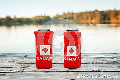 Two cans of beer in red cozy beer can cooler with Canadian flag standing on wooden pier by lake outdoors. Friends celebrating Stock Photo