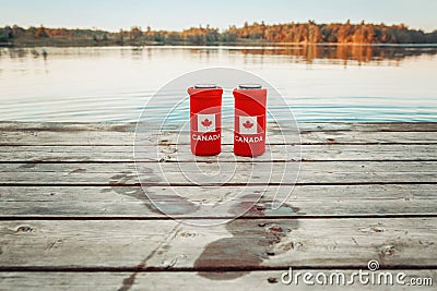 Two cans of beer in red cozy beer can coolers with Canadian flag standing on wooden pier by lake outdoor. Wet footprints on wooden Stock Photo