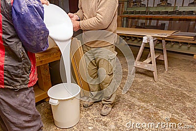 Two carpentry workers pour wood glue from a large plastic bucket into another empty Stock Photo