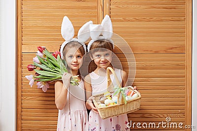 Two charming little sisters in the dresses with white rabbit`s ears on their heads holds flowers and a basket with the Stock Photo