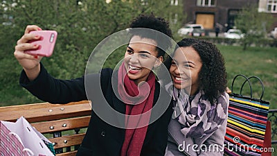 Two cheerful mixed race woman with shopping bags talking at video call with smartpone. Young girls sitting on bench have Stock Photo