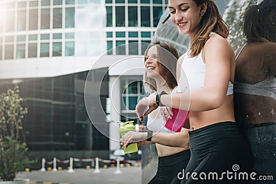 Two cheerful women runners stand leaning against trailer, rest after training, drink water, looking on pulsometer. Stock Photo