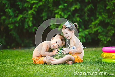 Two children, Caucasian brother and sister, sitting on green grass in backyard of house and hugging big tasty sweet watermelon Stock Photo