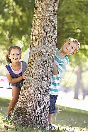 Two Children Hiding Behind Tree In Park Stock Photo