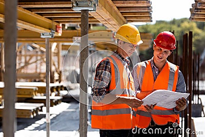 Two civil engineers dressed in orange work vests and helmets explore construction documentation on the building site Stock Photo