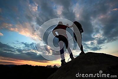Silhouettes of man and woman look at sunset on the top of rock. Hikers couple on dramatic sky at sunset. Back view. Stock Photo