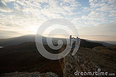 Two climbers standing on summit above clouds in the mountains holding hands. Silhouettes of hikers celebrating ascent on Stock Photo