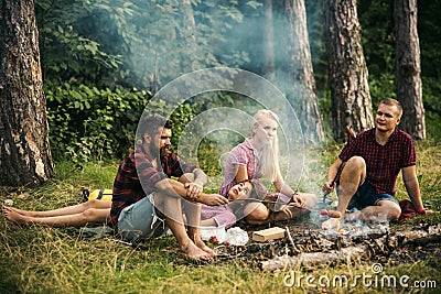 Two couples camping in woods. Men cooking sausages on campfire. Tired brunette lying on blond girls lap Stock Photo