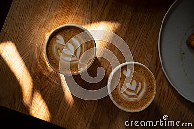 Two cups of fresh cappuccino with lovely latte art on brown wooden table with shadows and sun spots. Morning coffee for Stock Photo
