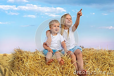 Two cute adorable caucasian siblings enjoy having fun sitting on top over golden hay bale on wheat harvested field near Stock Photo