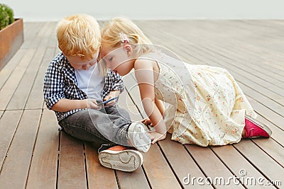 Two cute adorable white Caucasian toddlers boy and girl sitting together and playing games on cell mobile phone digital tablet. Stock Photo