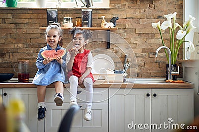 Two cute little girls sitting on kithcne countertop acting silly and enjoying healthy snack together Stock Photo