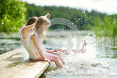 Two cute little girls sitting on a wooden platform by the river or lake dipping their feet in the water on warm summer day Stock Photo