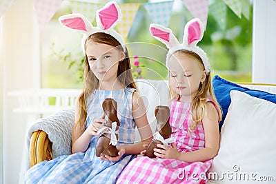 Two cute little sisters wearing bunny ears eating chocolate Easter rabbits. Kids playing egg hunt on Easter. Stock Photo