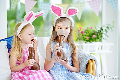 Two cute little sisters wearing bunny ears eating chocolate Easter rabbits. Kids playing egg hunt on Easter. Stock Photo