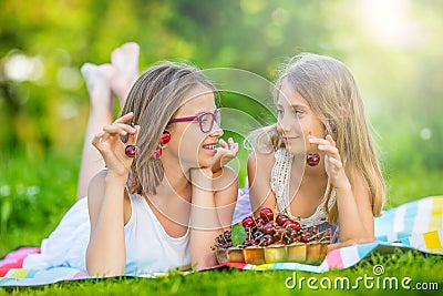 Two cute sisters or friends in a picnic garden lie on a deck and eat freshly picked cherries Stock Photo