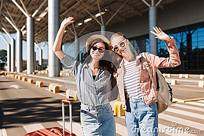 Two cute smiling girls in sunglasses happily looking in camera while raising hands up with suitcase and backpack on Stock Photo