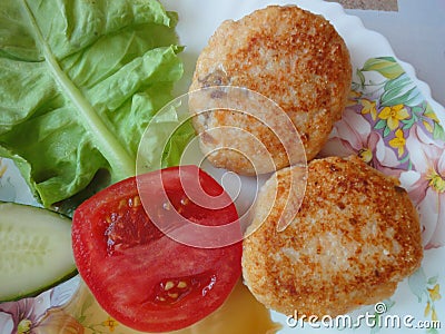 Two cutlets, a leaf of a lettuce, a tomato and a cucumber on a white plate / Ready lunch / Served dish with food / Stock Photo