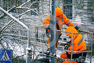 Two electricians in orange overalls on a lift work on top of an electric pole with cable lines against the backdrop of Editorial Stock Photo