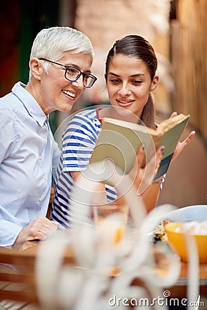 Two female friends of different generations reading a book together while they have a drink in the bar. Leisure, bar, friendship, Stock Photo