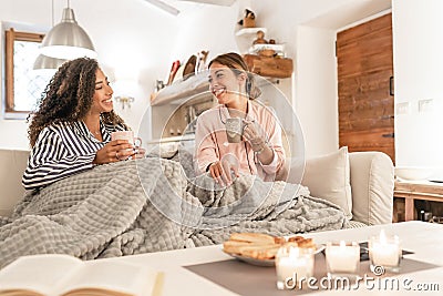 Two fine young women sitting on the sofa with a blanket on their legs laughing enjoying the house in winter having tea with Stock Photo