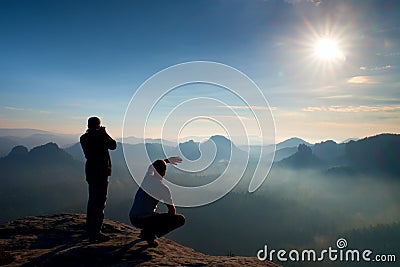 Two friends. Hiker thinking and photo enthusiast takes photos stay on cliff. Dreamy fogy landscape, blue misty sunrise in a beaut Stock Photo