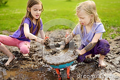 Two funny little girls playing in a large wet mud puddle on sunny summer day. Children getting dirty while digging in muddy soil Stock Photo