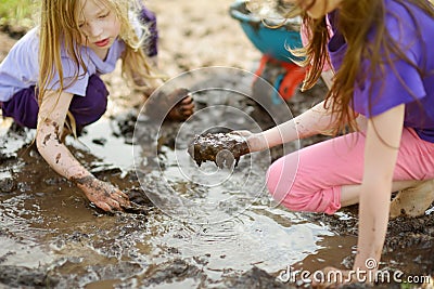 Two funny little girls playing in a large wet mud puddle on sunny summer day. Children getting dirty while digging in muddy soil. Stock Photo