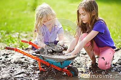 Two funny little girls playing in a large wet mud puddle on sunny summer day. Children getting dirty while digging in muddy soil. Stock Photo