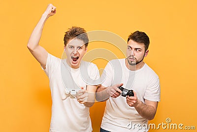 Two gamers in white T-shirts hold the joystick in their hands and emotionally play video games, isolated on a yellow background. Stock Photo