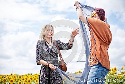 Two girlfriends, wearing boho hippie clothes, holding grey transparent shawl scarf, smiling, laughing in the middle of yellow Stock Photo