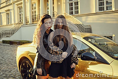 Two girls on a background of a yellow car. Beautiful brunettes near the cottage arrived on an expensive car. Famous girls. Stock Photo