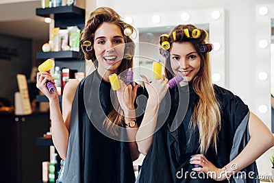 Two girls curling hair with curlers wearing cape having fun laughing putting rollers on fingers in hairdressing salon Stock Photo