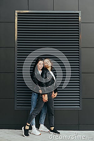 Two girls in dark casual clothes stand on a black background, hugging and posing at the camera.Attractive best girlfriends are Stock Photo