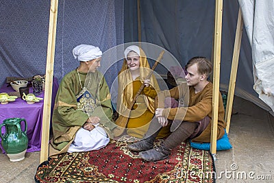 two girls and a guy in bright ancient historical dresses sit on a red Turkish carpet in a marquee. Editorial Stock Photo