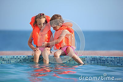 Two girls in lifejackets sitting on ledge pool Stock Photo