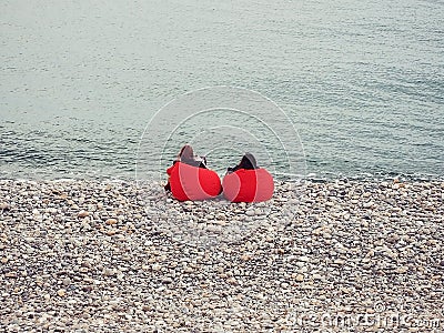 Two girls sit in red armchair bags on the pebble sea shore on a cool spring day Stock Photo