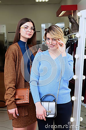 Two girls try on glasses in front of a large mirror in the store. Girls with glasses. Season of sales and discounts. Save money Stock Photo