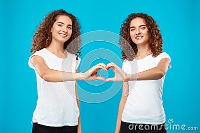 Two girls twins showing heart with hands over blue background. Stock Photo